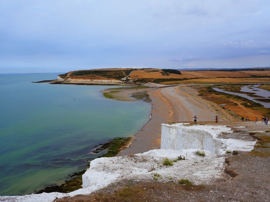 The image depicts a scenic coastal landscape with towering white chalk cliffs extending along the shoreline in the background, under a partly cloudy sky. In the foreground, a dirt pathway leads downhi