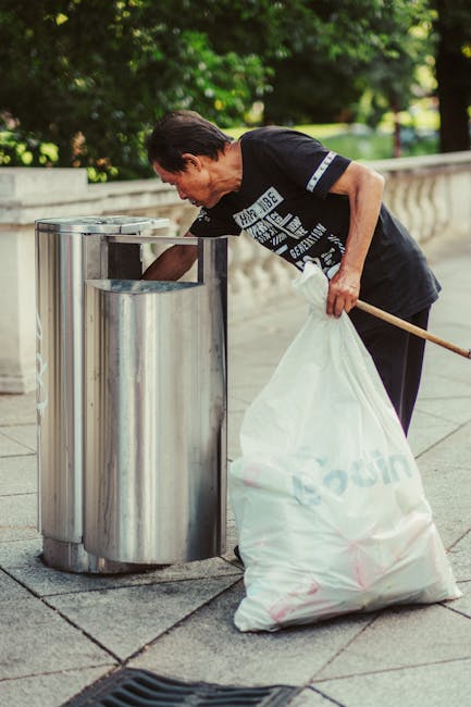 A middle-aged man wearing a black T-shirt with white printed text and black pants is seen disposing of a large white rubbish bag into a metal public litter bin alongside a concrete pathway. The bin ha