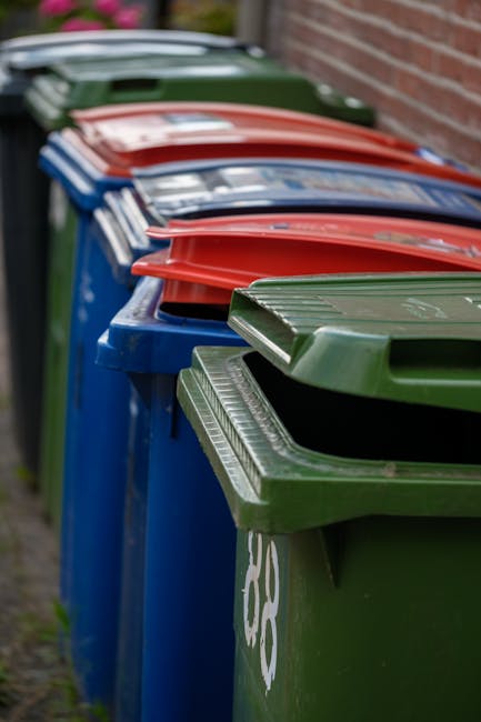 A middle-aged man wearing a black T-shirt with white printed text and black pants is seen disposing of a large white rubbish bag into a metal public litter bin alongside a concrete pathway. The bin ha