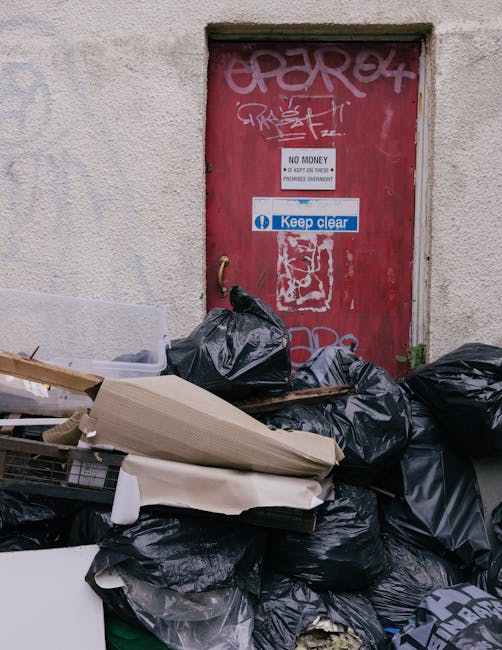 Two large black plastic rubbish bags, slightly crumpled and tied at the top, are positioned on the pavement next to a dark metal fence with vertical bars. The bags appear to contain waste materials an