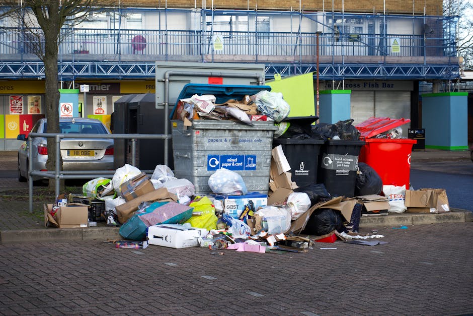 A large pile of mixed household waste and rubbish bags is gathered on a paved sidewalk in front of a row of commercial units and a parked silver car, with some cardboard boxes and loose paper scattere